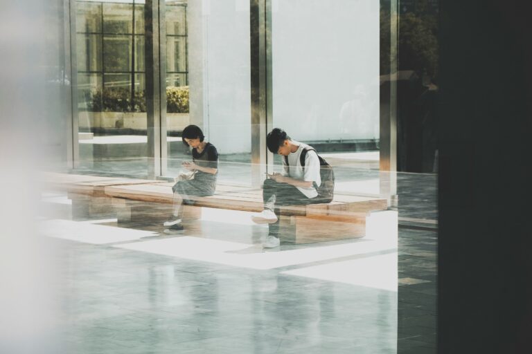 A photo of two people sitting on a long wooden bench in an open hallway with high windows.